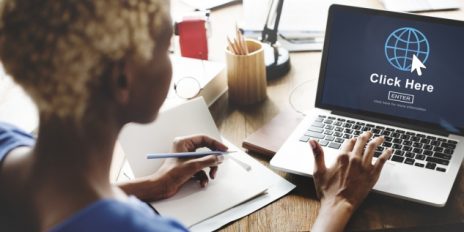 Ein Frau an einem Schreibtisch mit einen Laptop auf dem steht "Click Here".
