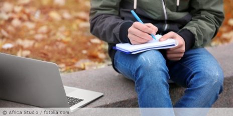 Ein Mann auf einer Mauer mit Stift und Block in der Hand, daneben ein Laptop.