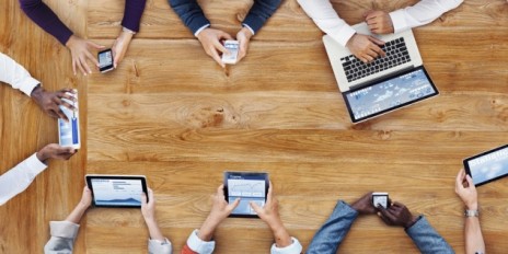 Personen sitzen zusammen an einem Tisch und schauen sich etwas auf dem Smartphone, Ipad oder Laptop an.