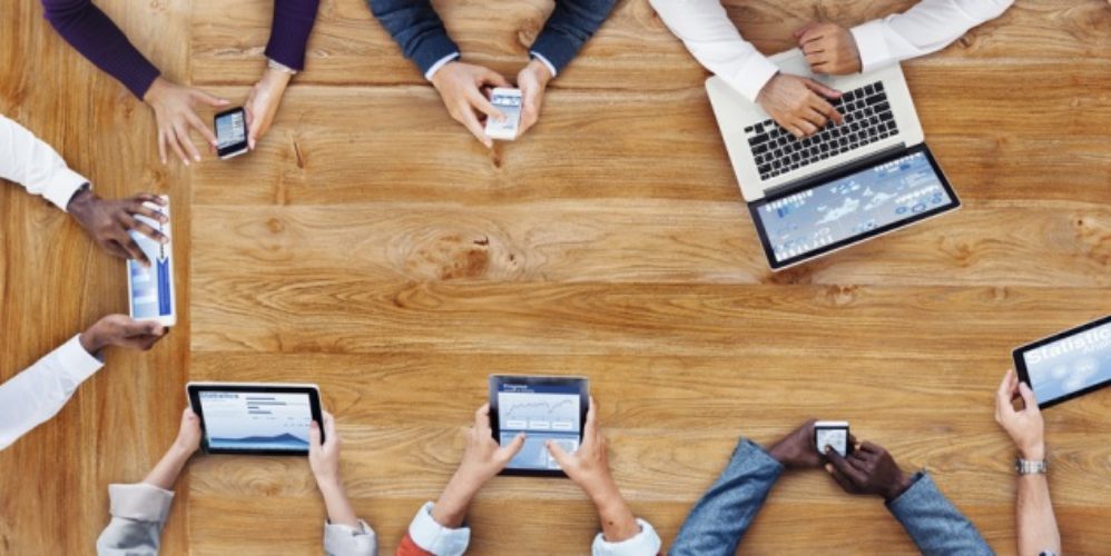 Personen sitzen zusammen an einem Tisch und schauen sich etwas auf dem Smartphone, Ipad oder Laptop an.