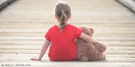 Kind sitzt mit Teddybär auf dem Boden.