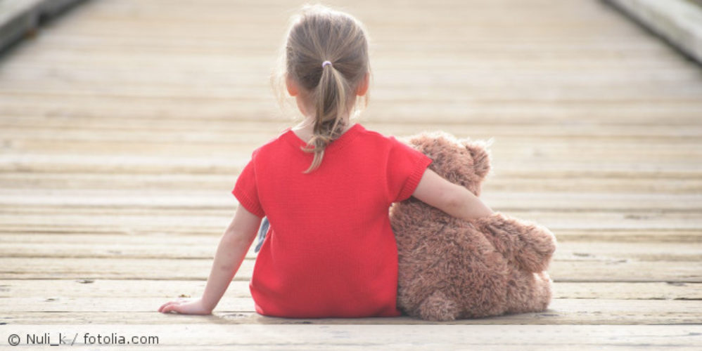 Kind sitzt mit Teddybär auf dem Boden.