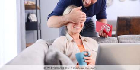 Mann hält einen Gutschein in der Hand und seiner Partnerin die Augen zu als Überraschung.