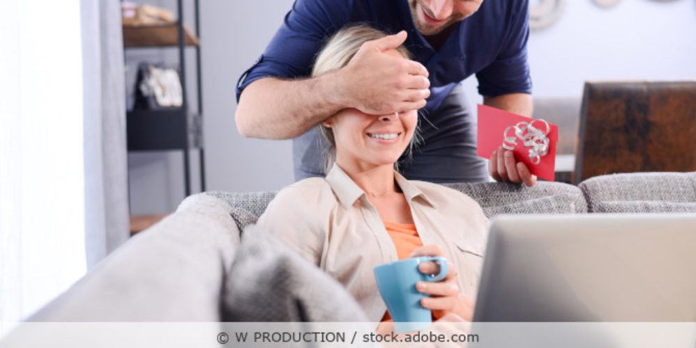 Mann hält einen Gutschein in der Hand und seiner Partnerin die Augen zu als Überraschung.