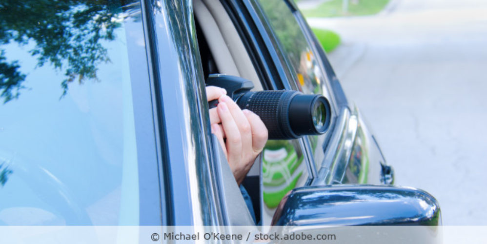 Ein Detektiv mit Kamera sitzt im Auto und fotografiert durch das geöffnete Autofenster.