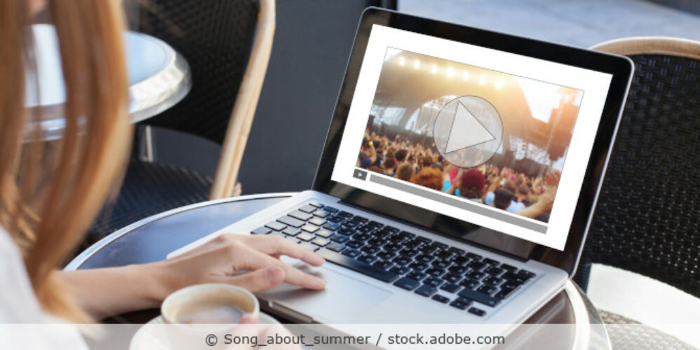 Frau sitzt mit einem Kaffee vor ihrem Laptop und schaut ein Video von einem Konzert.