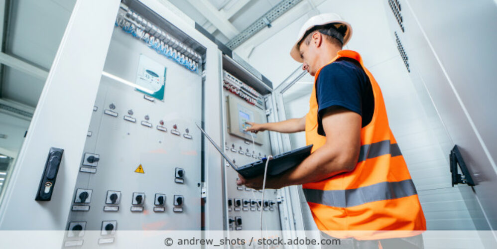 Arbeiter steht vor einem geöffneten Schaltkasten mit einem Laptop in der Hand.