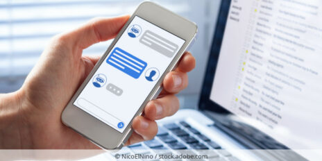 A hand holding a smartphone that shows the conversation with a chatbot and a laptop-screen in the background. Eine Hand hält ein Smartphone zur Konversation mit einem Chatbot, im Hintergrund ist ein geöffneter Laptop.