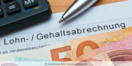 Eine Lohn- und Gehaltsabrechnung liegt auf einem Holztisch, verdeckt von ein paar Euro-Geldscheinen und zusammen mit einem Taschenrechner und einem Stift.