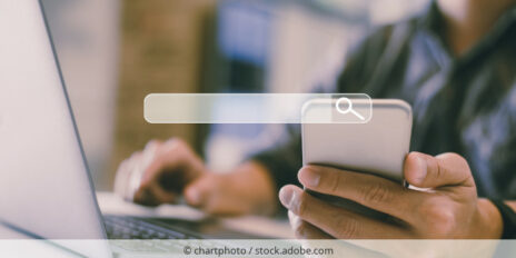 Person sitzt am Laptop und nutzt Suchfunktion eines Smarphones.