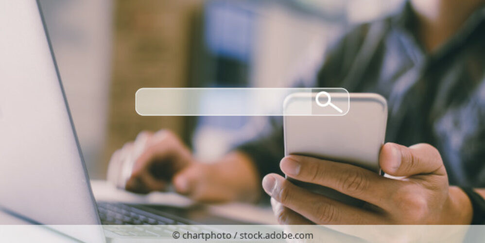 Person sitzt am Laptop und nutzt Suchfunktion eines Smarphones.