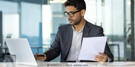 Mann sitzt in einem Büro an seinem Laptop, hält ein Dokument in der Hand und schaut auf den Bildschirm.