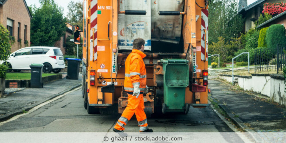 Stehendes Müllfahrzeug auf einer Straße in einem Wohngebiet, ein Müllwerker leert gerade eine grüne Tonne.