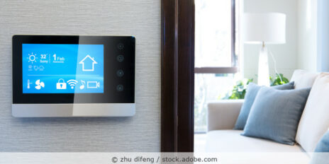 Touchscreen für Smart-Home-Steuerung hängt an einer Wand, im Hintergrund ist ein Wohnzimmer zu sehen.