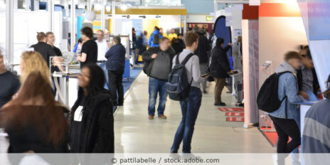 Gang auf einer Fachmesse mit Messeständen links und rechts und Besuchern, die sich informieren.