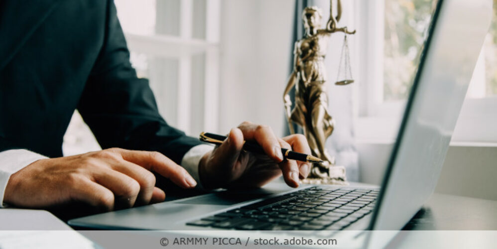 Mann sitzt im Büro und arbeitet an einem Laptop; auf dem Schreibtisch steht eine Justitia-Figur.