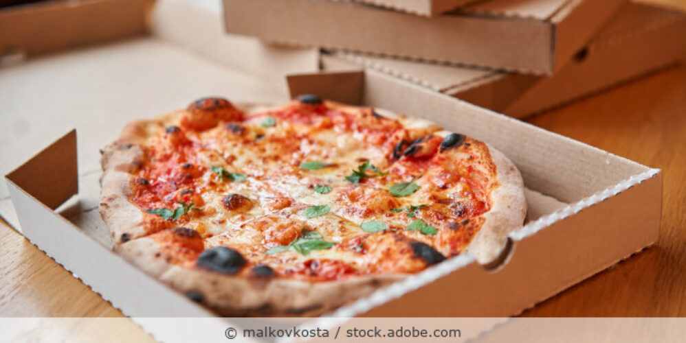 Eine Pizza Margherita liegt in einem geöffneten Pizzakarton auf einem Tisch mit anderen Pizzakartons.
