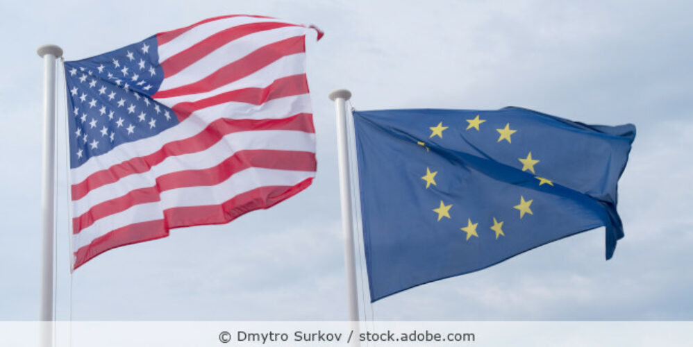 Die Flagge der USA und die Europaflagge wehen vor blauem Himmel.