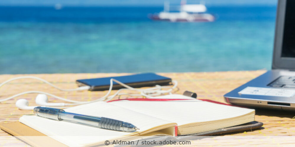 Ein Notizbuch, ein Handy und ein Laptop liegen auf einem Tisch, im Hintergrund ist ein Boot auf türkisblauem Wasser zu sehen.