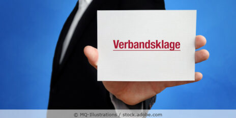 Ein Mann im schwarzen Anzug hält ein weißes Schild in der Hand auf dem in roter Schrift das Wort Verbandsklage steht.