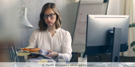 Mitarbeiterin sitzt im Büro an ihrem Schreibtisch und schaut skeptisch auf einen Berg an Unterlagen.
