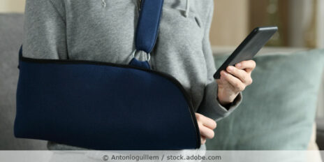 Eine Frau hat ihren Arm in einer dunkelblauen Armschlinge und hält in der anderen Hand das Handy.