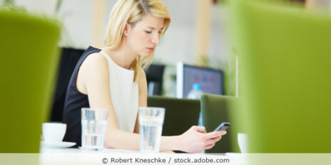 Eine blonde Frau im Business-Outfit ist an einem Besprechungstisch und schaut auf ihr Handy.