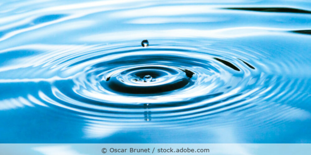 Ein Tropfen fällt auf eine glatte Wasseroberfläche und erzeugt Ringe bzw. kleine Wellen.