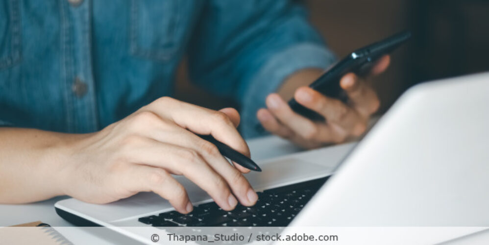Ein Mann arbeitet an einem Laptop und hält sein Handy in einer Hand.
