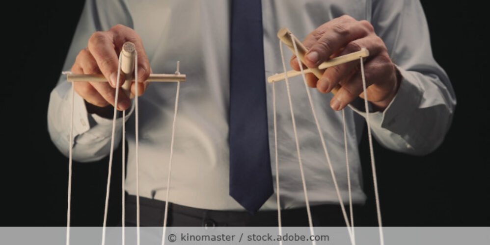 Ein Geschäftsmann in grauem Hemd und schwarzer Krawatte steuert eine Marionette mit einem hölzernen Greifer und Fäden.
